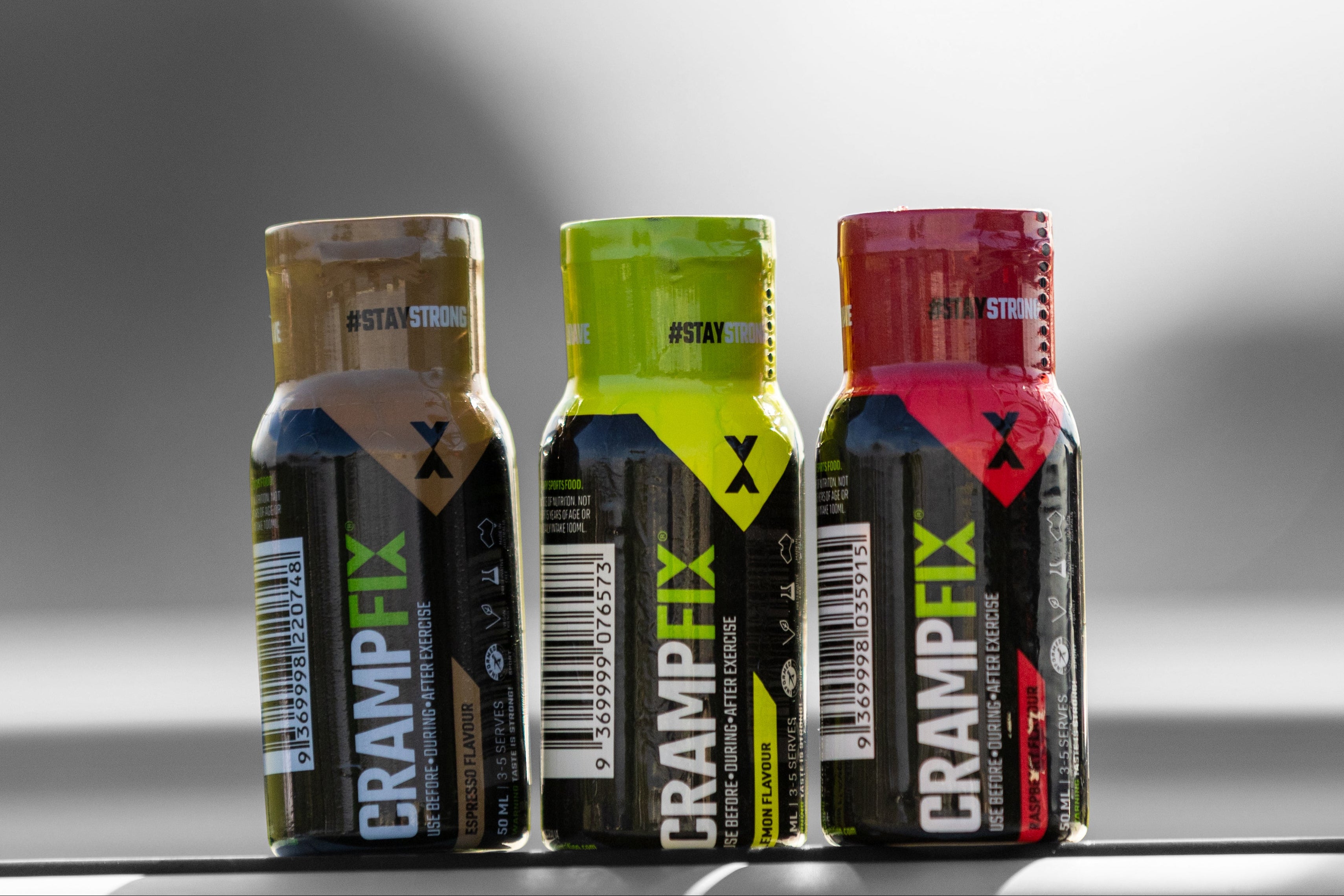 Three CrampFix 50ml bottles in different colors (black with gold, black with lime green, and black with red) lined up on a reflective surface against a gray background.